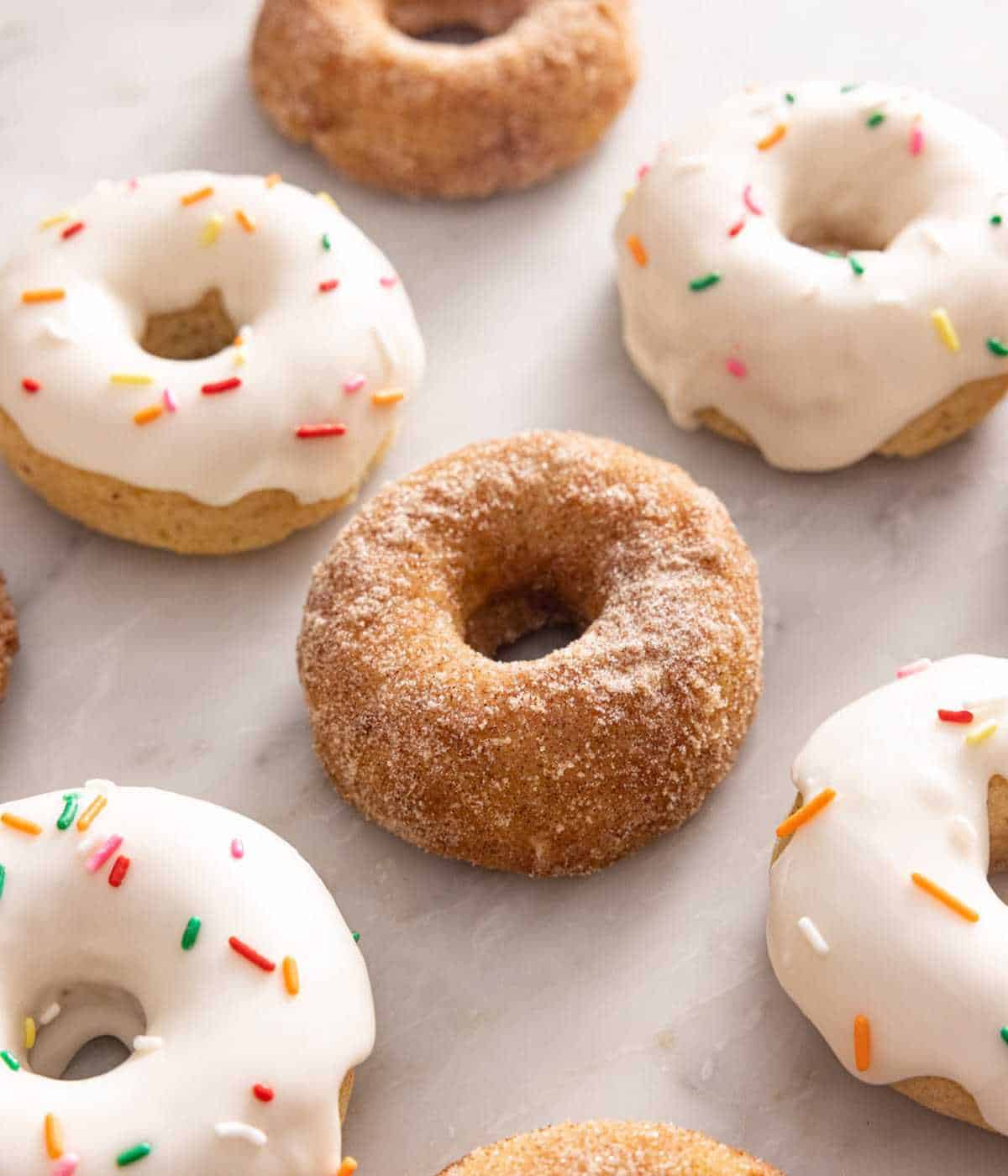 Baked donuts on a marble surface, some coated with glaze, others with cinnamon sugar.