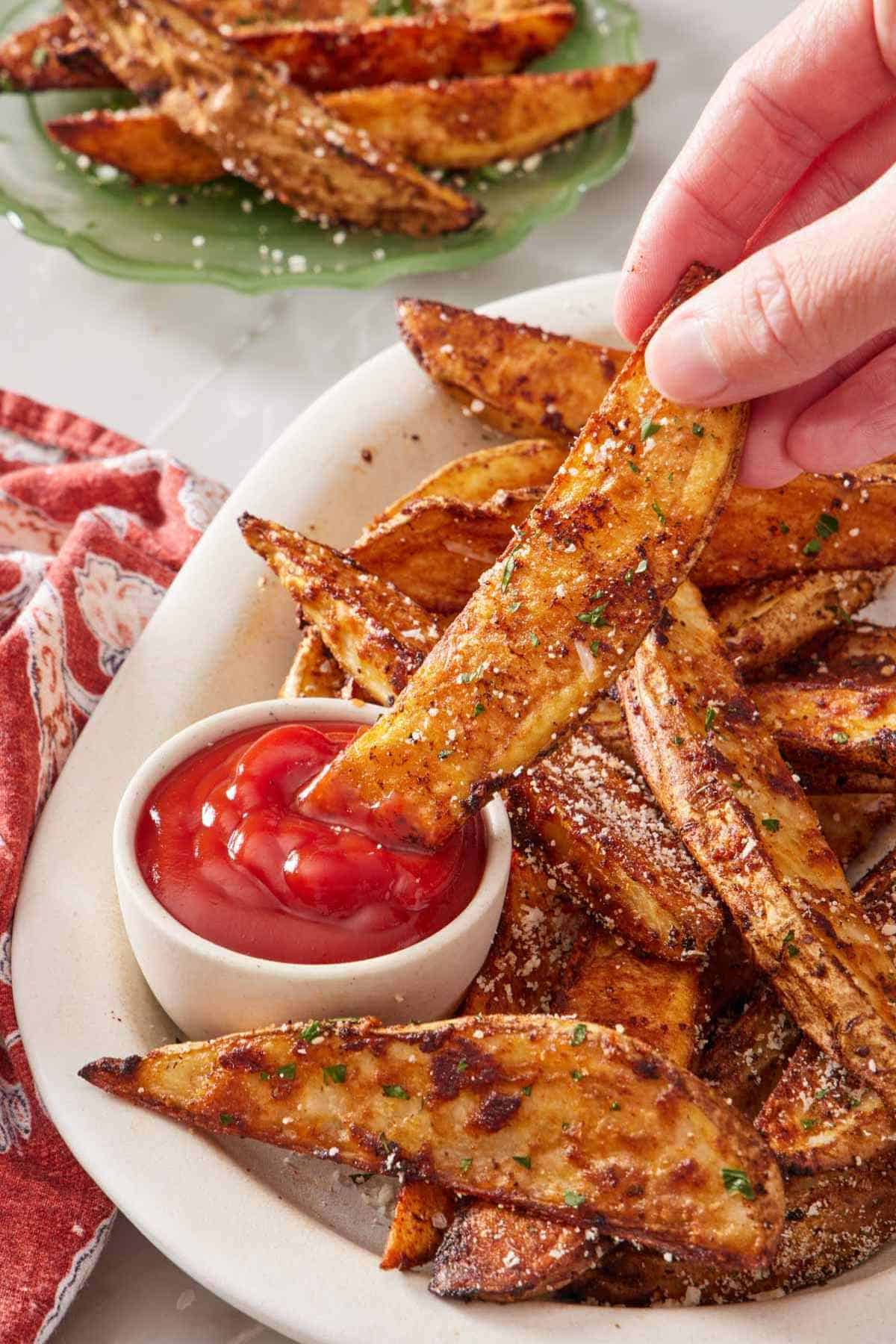 A plate of oven-baked potato wedges, one being dipped into a small bowl of ketchup.