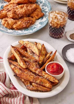 Baked potato wedges on a serving platter with a small dish of ketchup, with fried chicken and two glasses of soda.