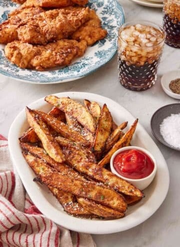 Baked potato wedges on a serving platter with a small dish of ketchup, with fried chicken and two glasses of soda.