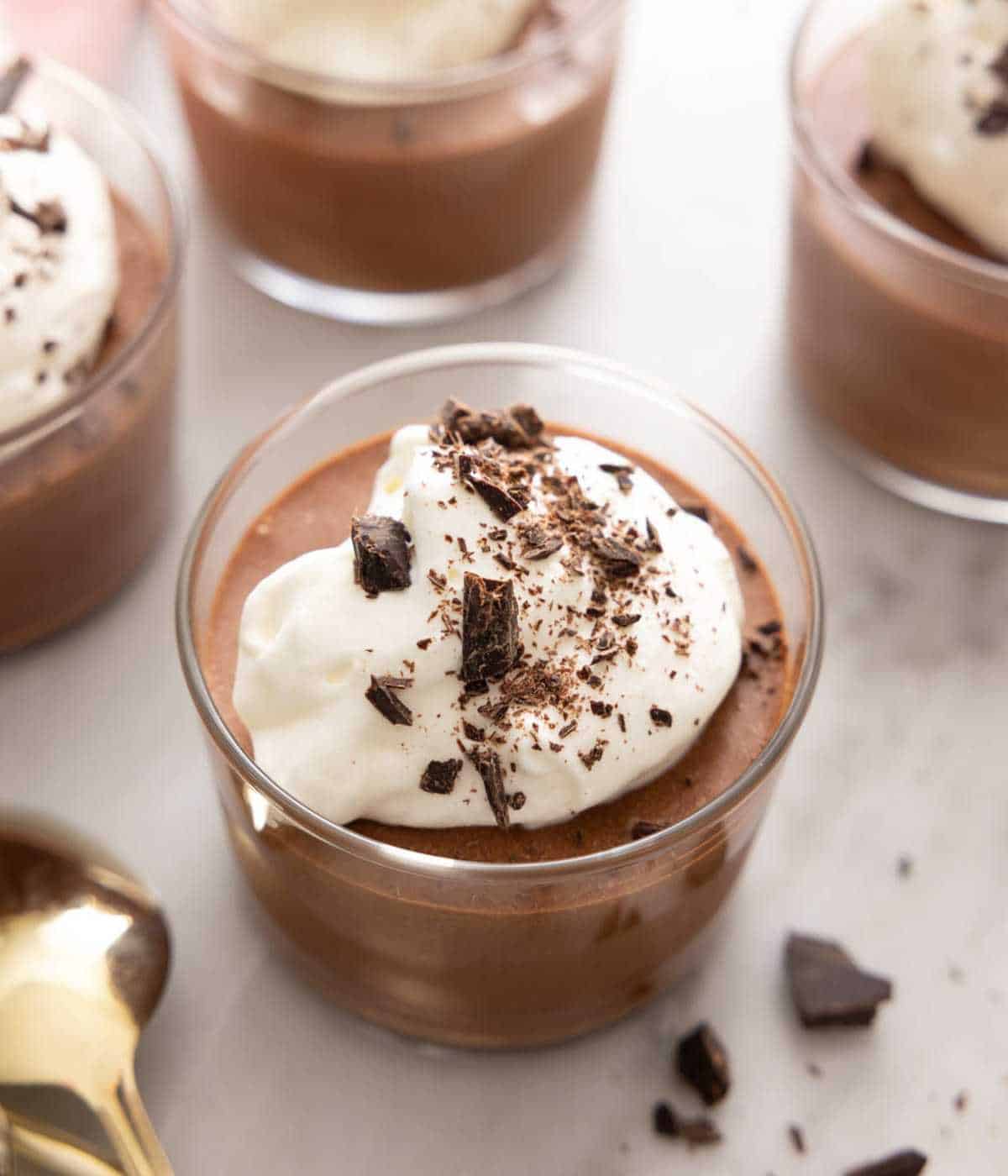 A few glasses of chocolate mousse, with one in focus, topped with whipped cream and chocolate shavings.