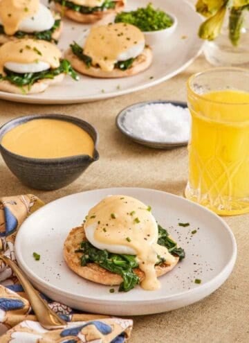 Eggs Florentine on a white plate next to a glass of juice, with hollandaise sauce, salt, and more eggs Florentine in the background.