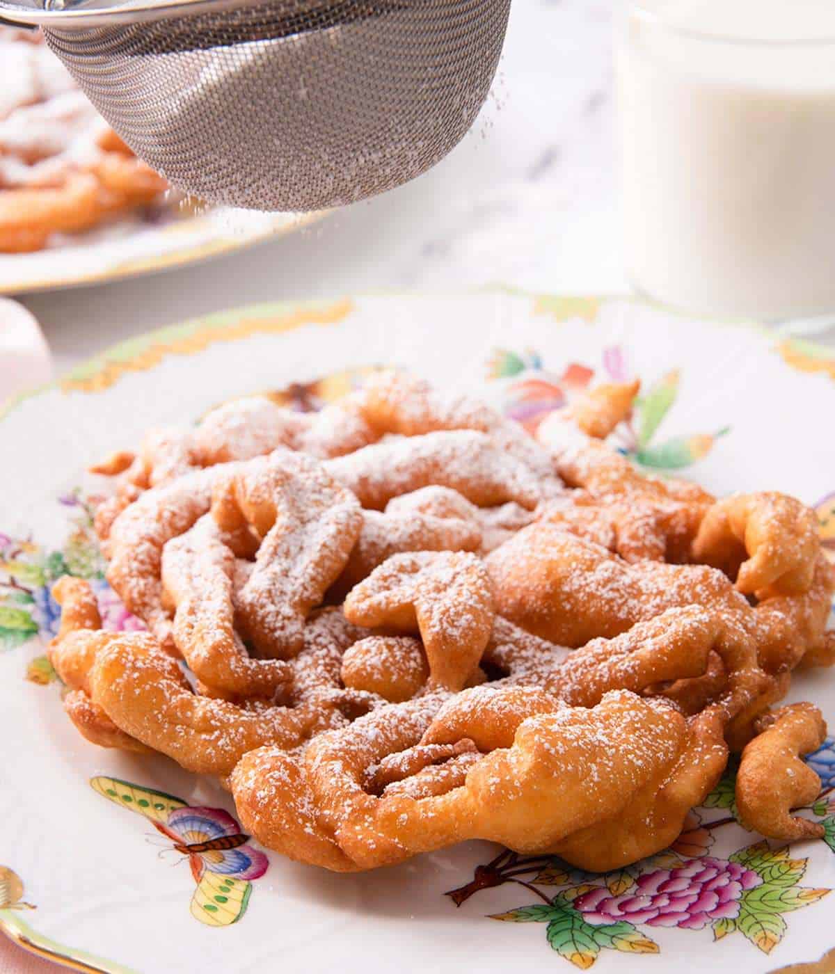 A funnel cake on a patterned plate being dusted with powdered sugar.