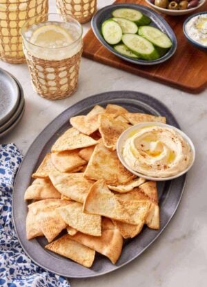 Homemade pita chips on a platter with hummus, with sliced cucumber, olives, and tzatziki in the background. A few drinks are next to the platter.