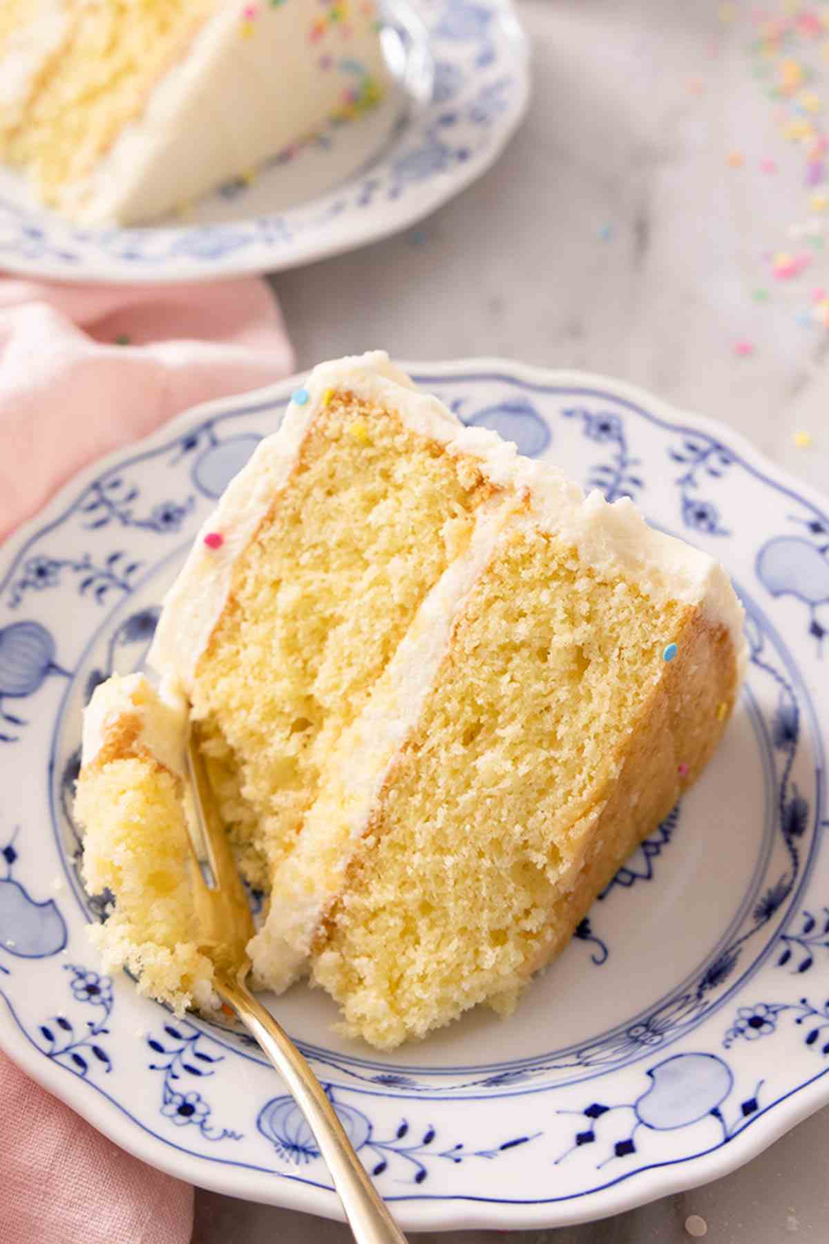 A slice of vanilla cake on a blue and white plate with a fork scooping some cake.