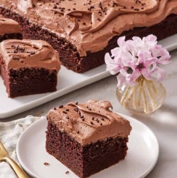 A piece of chocolate sheet cake on a small plate next to a small vase of flowers. The rest of the cake out of focus in the background.