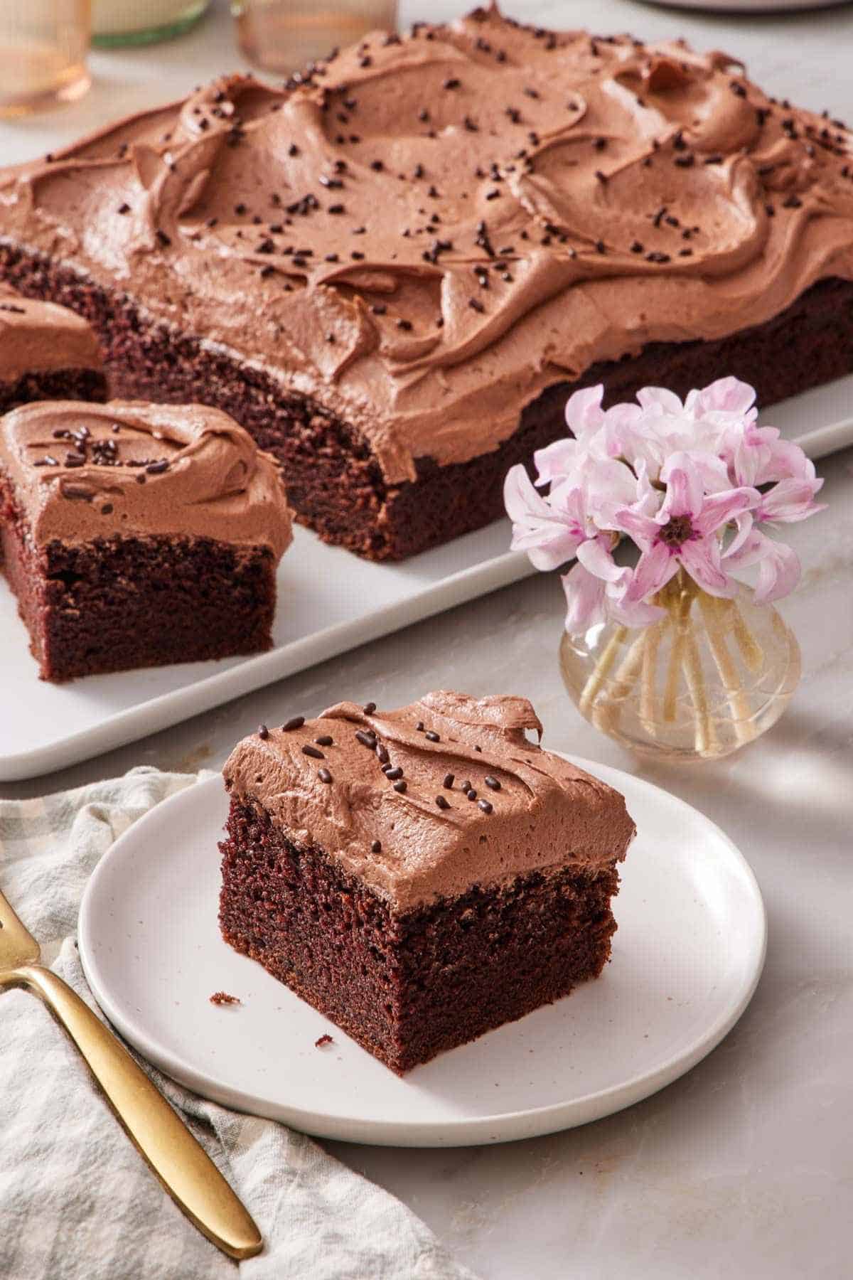 A piece of chocolate sheet cake on a small plate next to a small vase of flowers. The rest of the cake out of focus in the background.