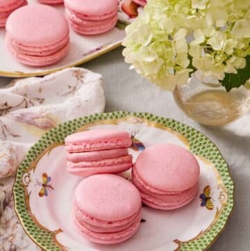 Three macarons on a plate next to a vase of flowers. A platter with more macarons in the background.