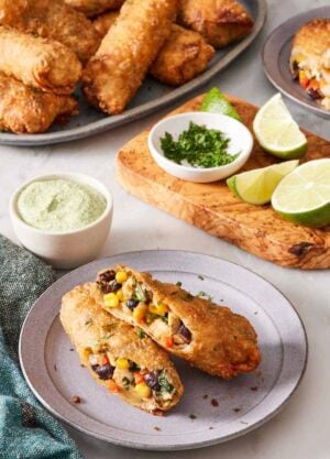 A southwest egg roll on a plate, halved to showcase the filling. Limes, dipping sauce, and more egg rolls in the background.