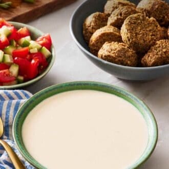 Pinterest graphic showing a bowl of tahini sauce with falafel and tomato and cucumber salad in the background.