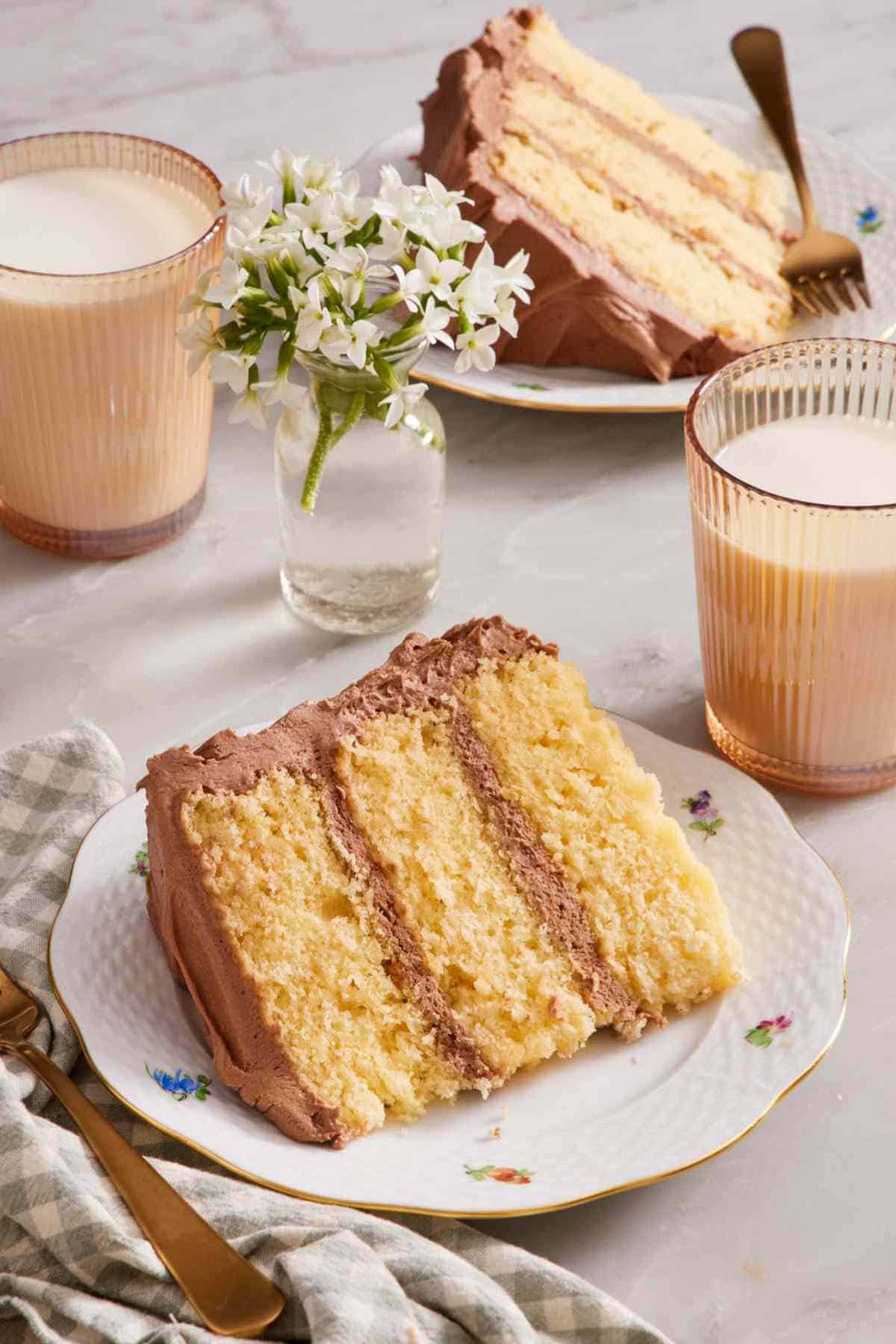 A slice of yellow cake on a small plate with glasses of milk and a vase of flowers.