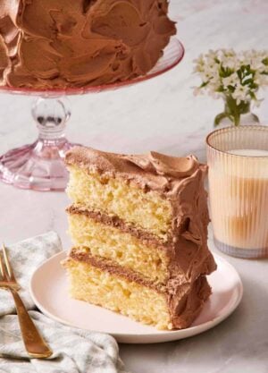 A slice of yellow cake standing up on a small plate alongside a glass of milk.