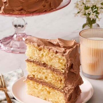 A slice of yellow cake standing up on a small plate alongside a glass of milk.