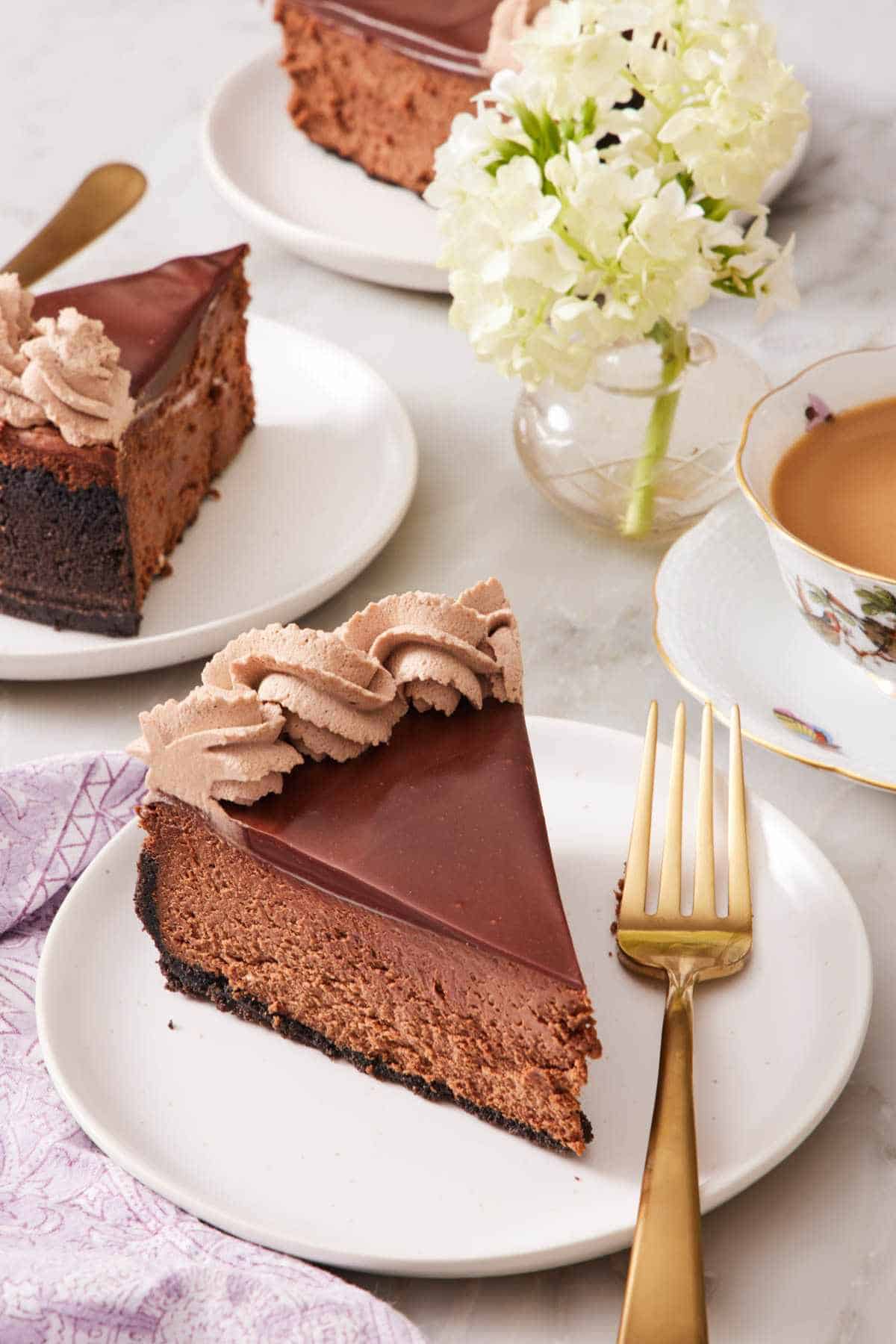 A slice of chocolate cheesecake on a plate with a fork with more slices almost out of frame behind it.