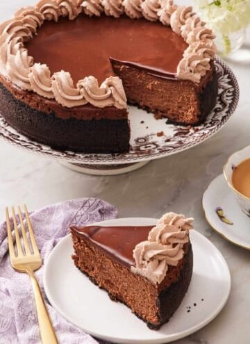 A slice of chocolate cheesecake on a small plate with the rest of the cheesecake on a cake stand right behind it.