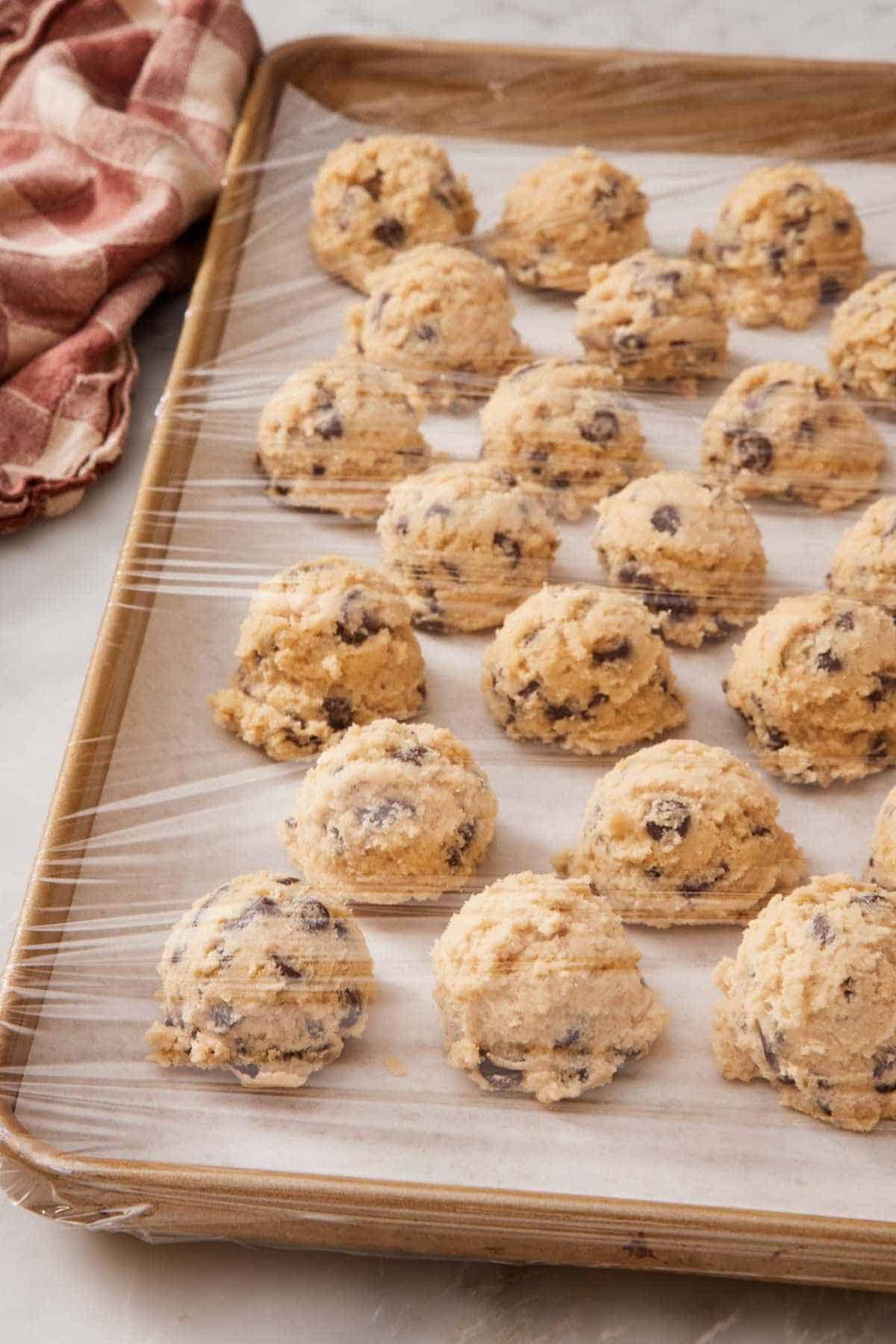 Cookie dough balls on a lined baking sheet, wrapped tightly with plastic wrap.