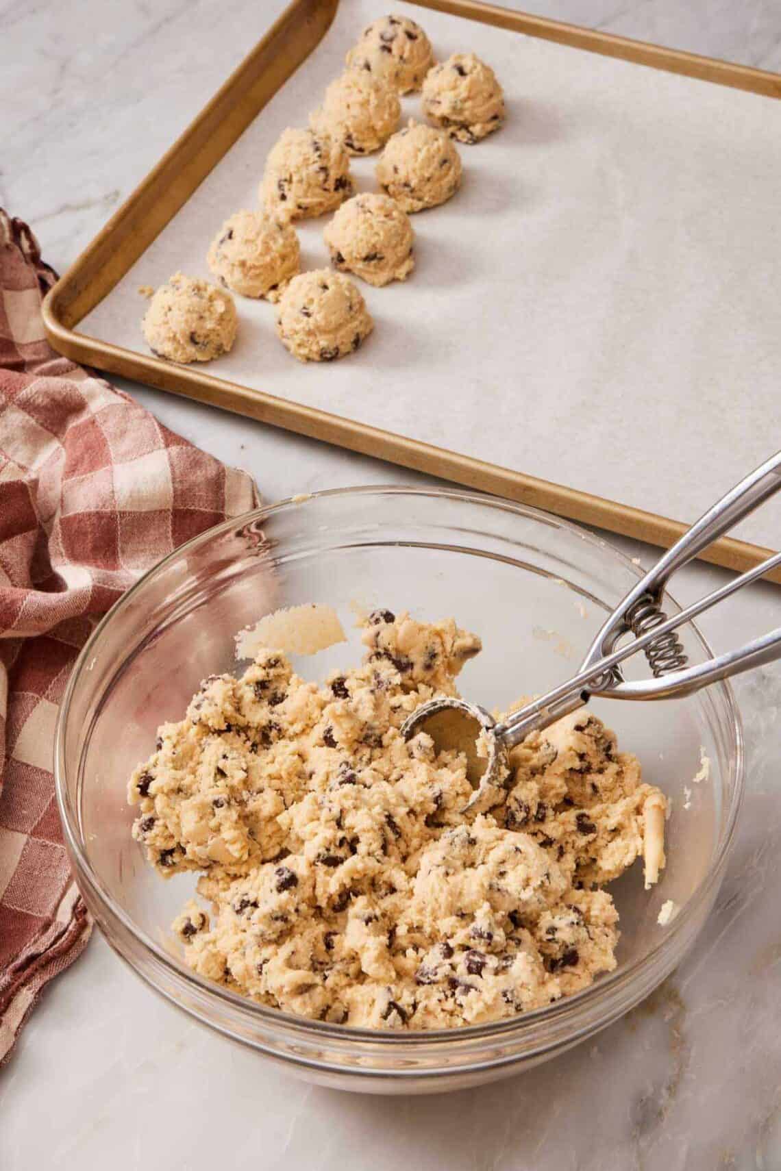 Cookie dough in a bowl with a cookie scoop, with scooped dough just behind on a baking sheet.