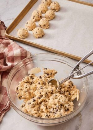 Cookie dough in a bowl with a cookie scoop, with scooped dough just behind on a baking sheet.