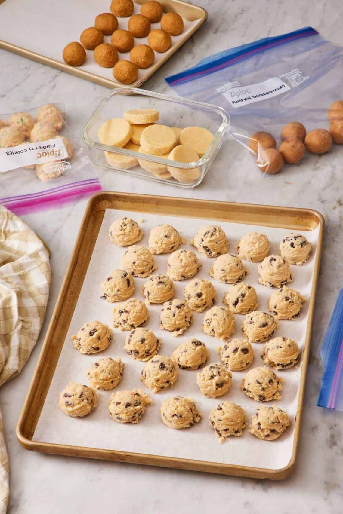 Cookie dough balls on a lined cookie sheet, some in a container, and others in freezer bags.