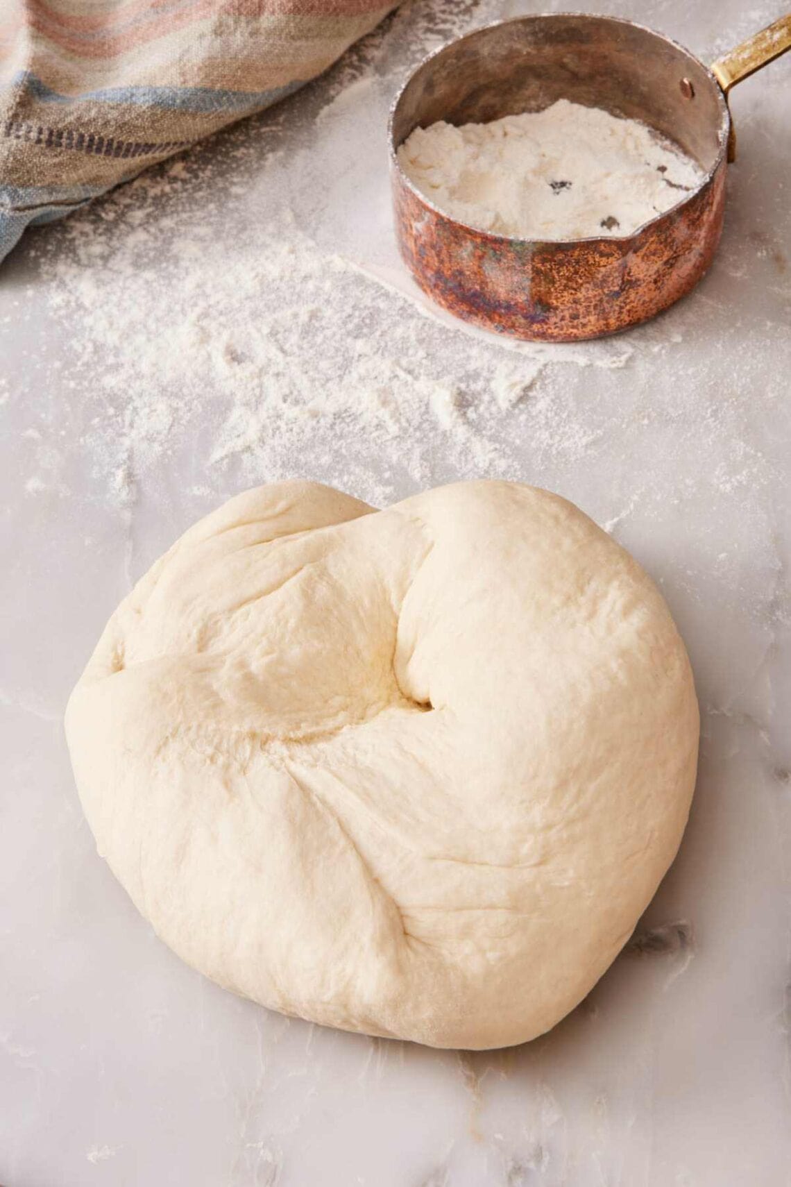 Kneaded dough on a countertop with flour sprinkled around, plus a measuring cup with more flour.