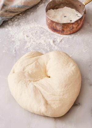 Kneaded dough on a countertop with flour sprinkled around, plus a measuring cup with more flour.