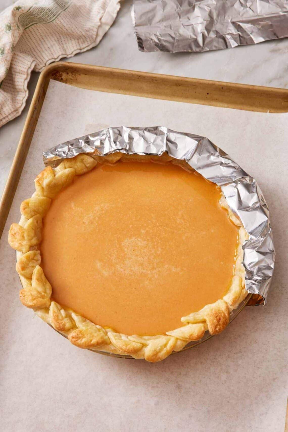 A pumpkin pie on a baking tray with half the edge shielded with foil.
