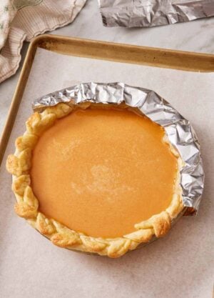 A pumpkin pie on a baking tray with half the edge shielded with foil.