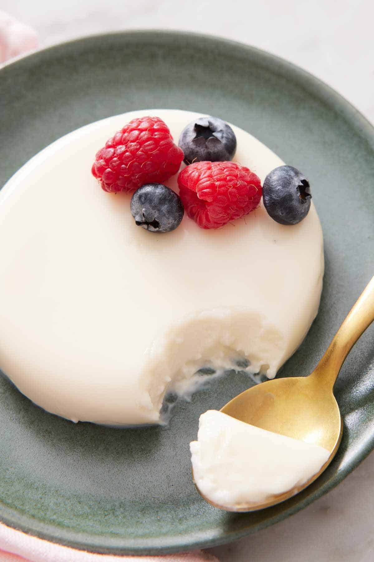 Panna cotta on a green plate with a little taken out with a spoon. It is topped with fresh berries.