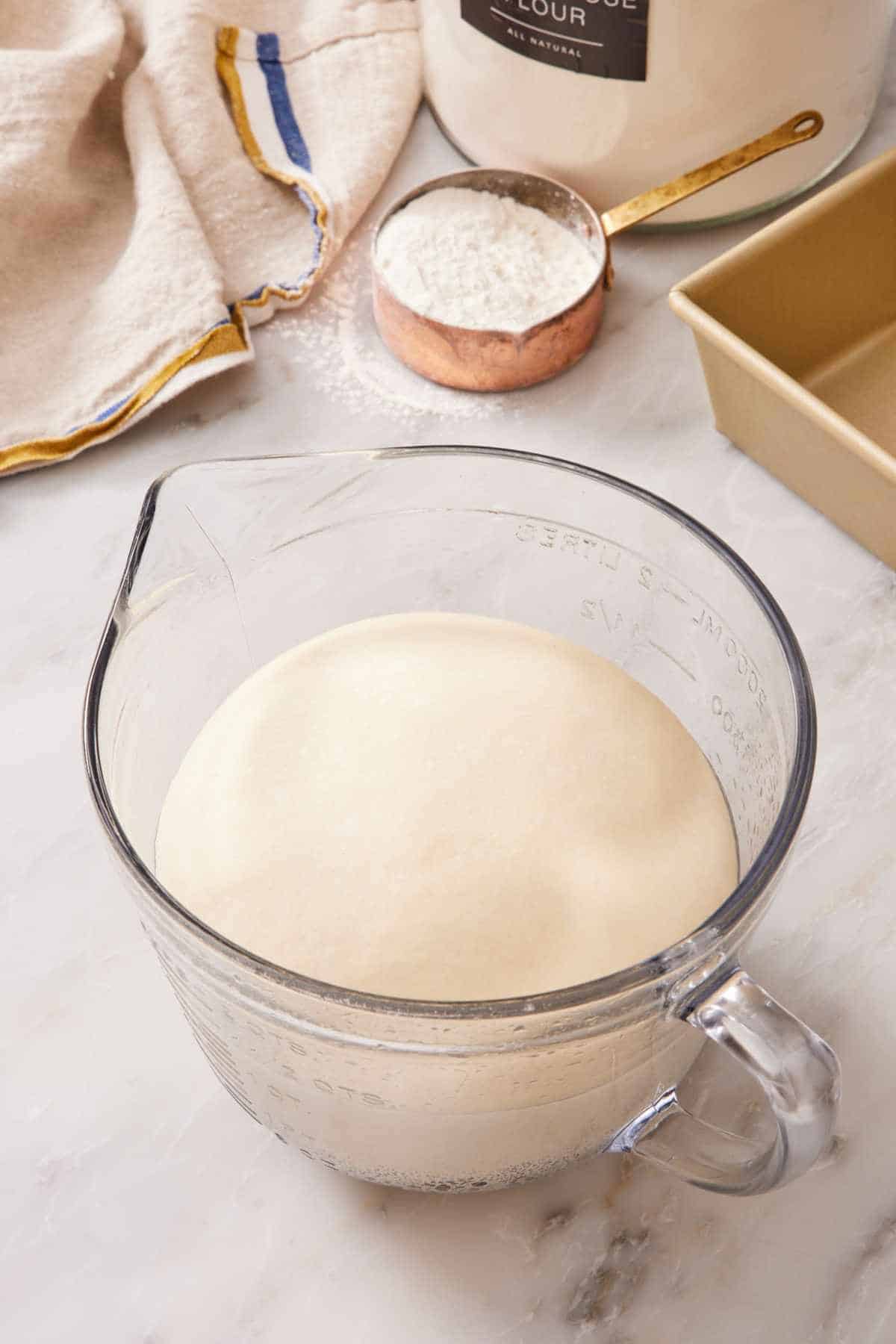 Bread dough proofing in a large measuring jug with a measuring cup of flour in the background.