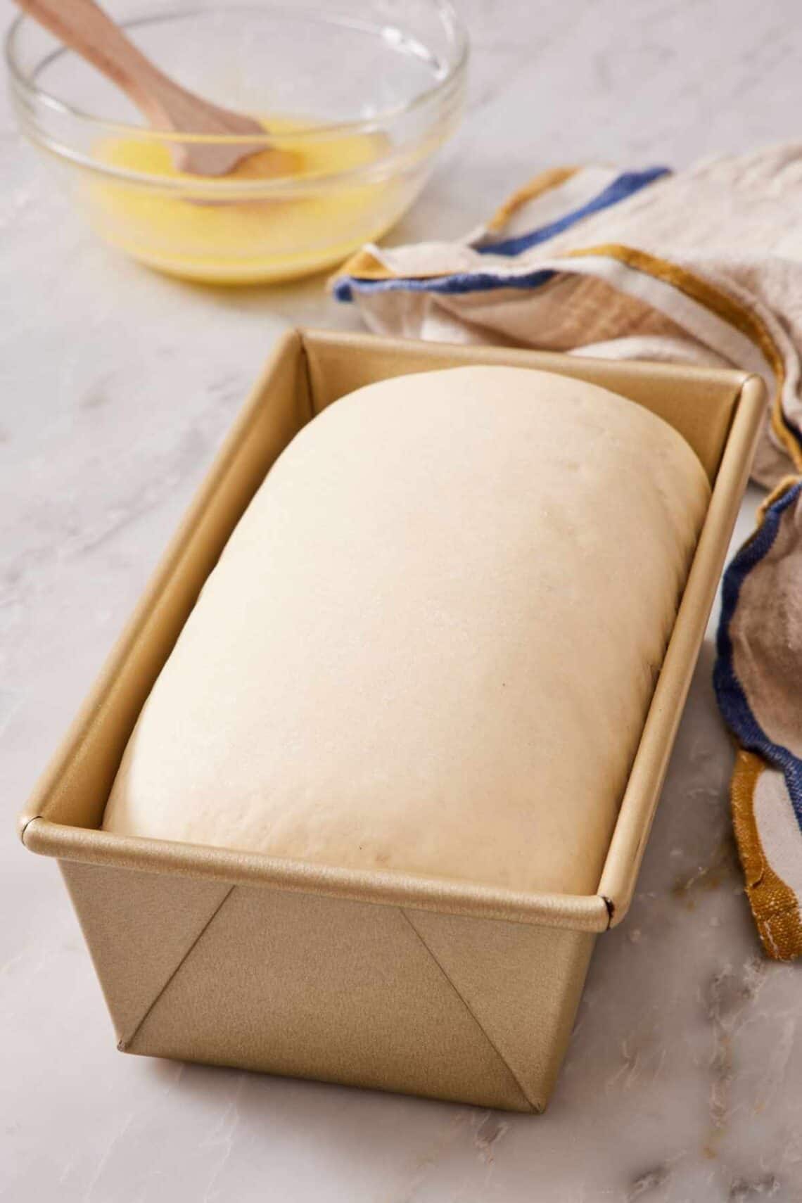 Proofed dough in a loaf pan with a bowl of melted butter out of focus in the background.