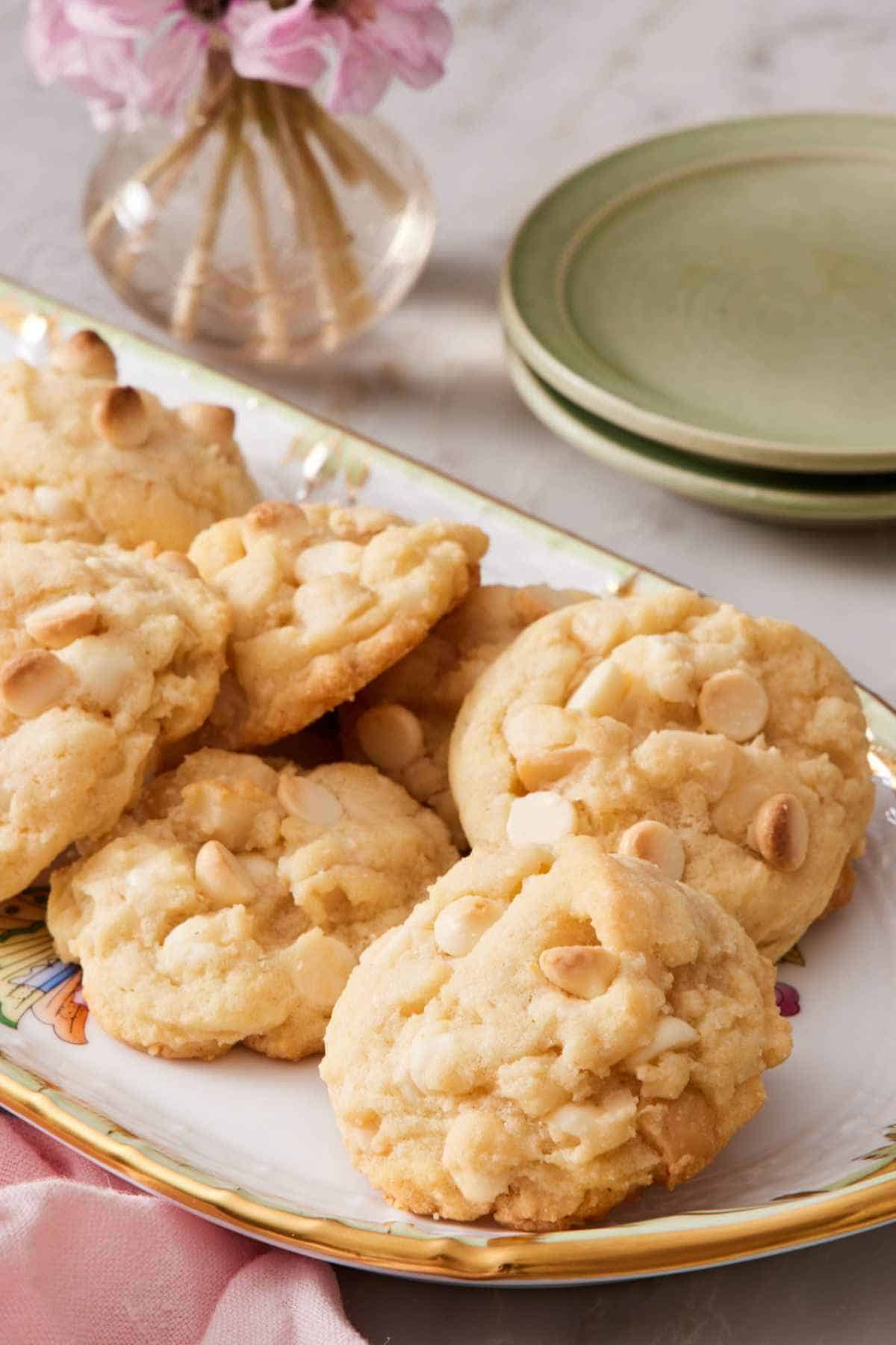 A platter of white chocolate macadamia nut cookies with small serving plates and a vase of flowers partly in frame.