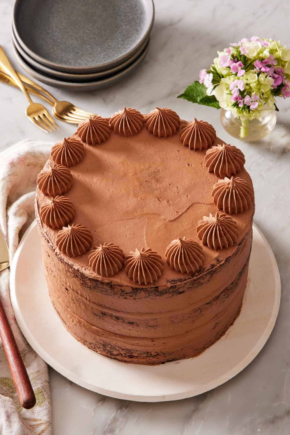 A whole, frosted chocolate zucchini cake on a white plate with small plates, forks, and flowers in the background.