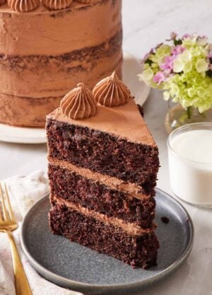A slice of chocolate zucchini cake on a small plate with a glass of milk on the side, and the rest of the cake almost out of frame in the background.