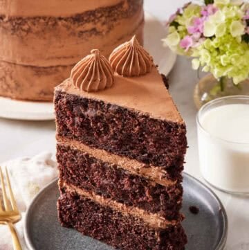 A slice of chocolate zucchini cake on a small plate with a glass of milk on the side, and the rest of the cake almost out of frame in the background.