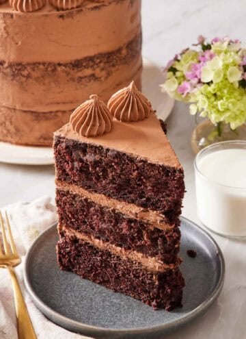 A slice of chocolate zucchini cake on a small plate with a glass of milk on the side, and the rest of the cake almost out of frame in the background.