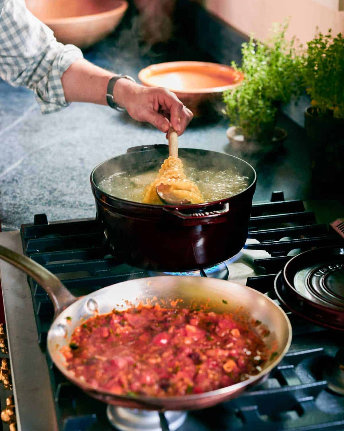 A spoon lifting pasta sheets out of a pot of boiling water, with red sauce in a pan in the foreground out of focus.