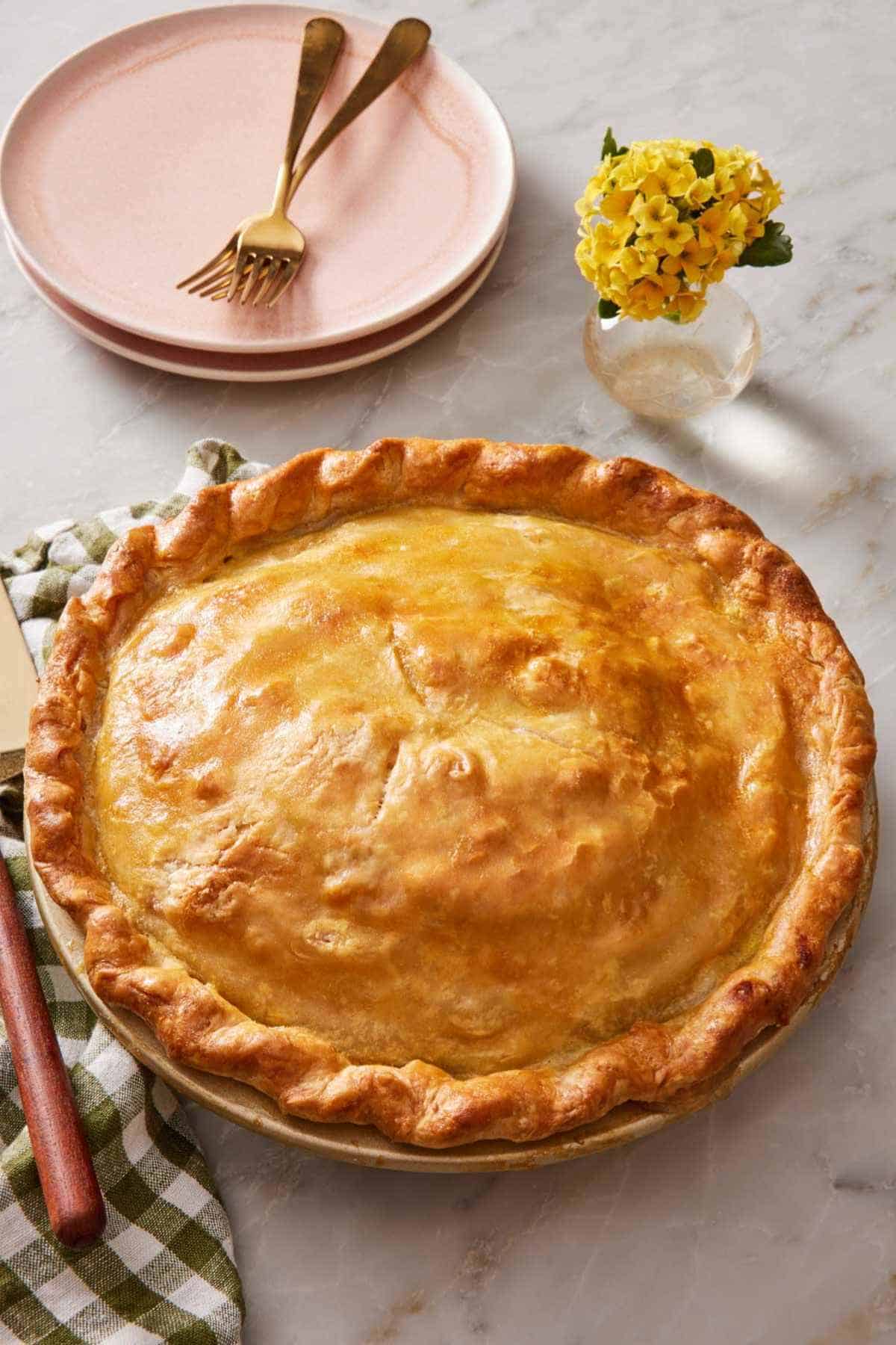 A baked double crust pie on a counter with a table linen next to it, and small plates behind it next to a small vase of flowers.