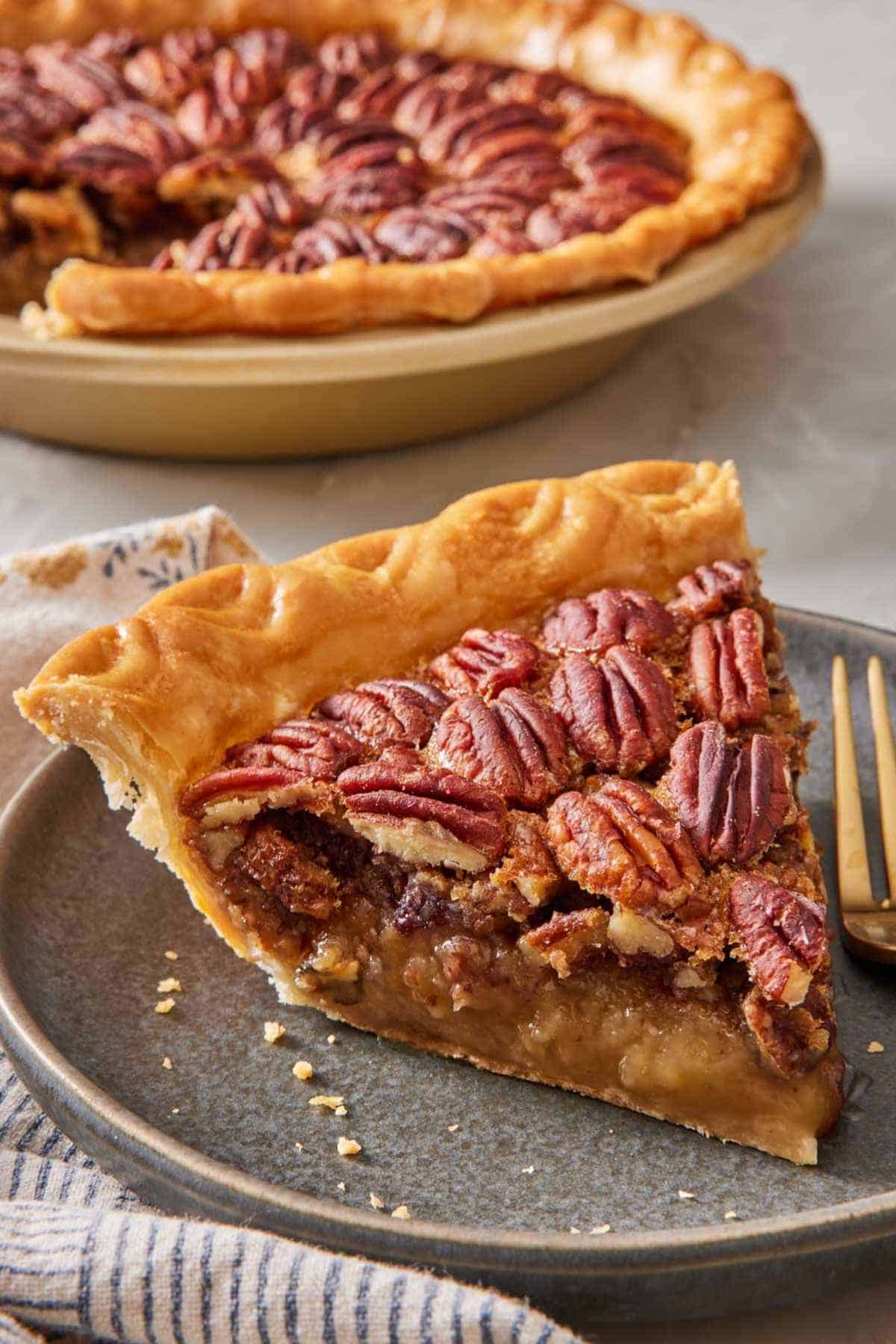 A close-up of a slice of pecan pie on a small plate with a fork. The rest of the pie is out of the focus in the background.