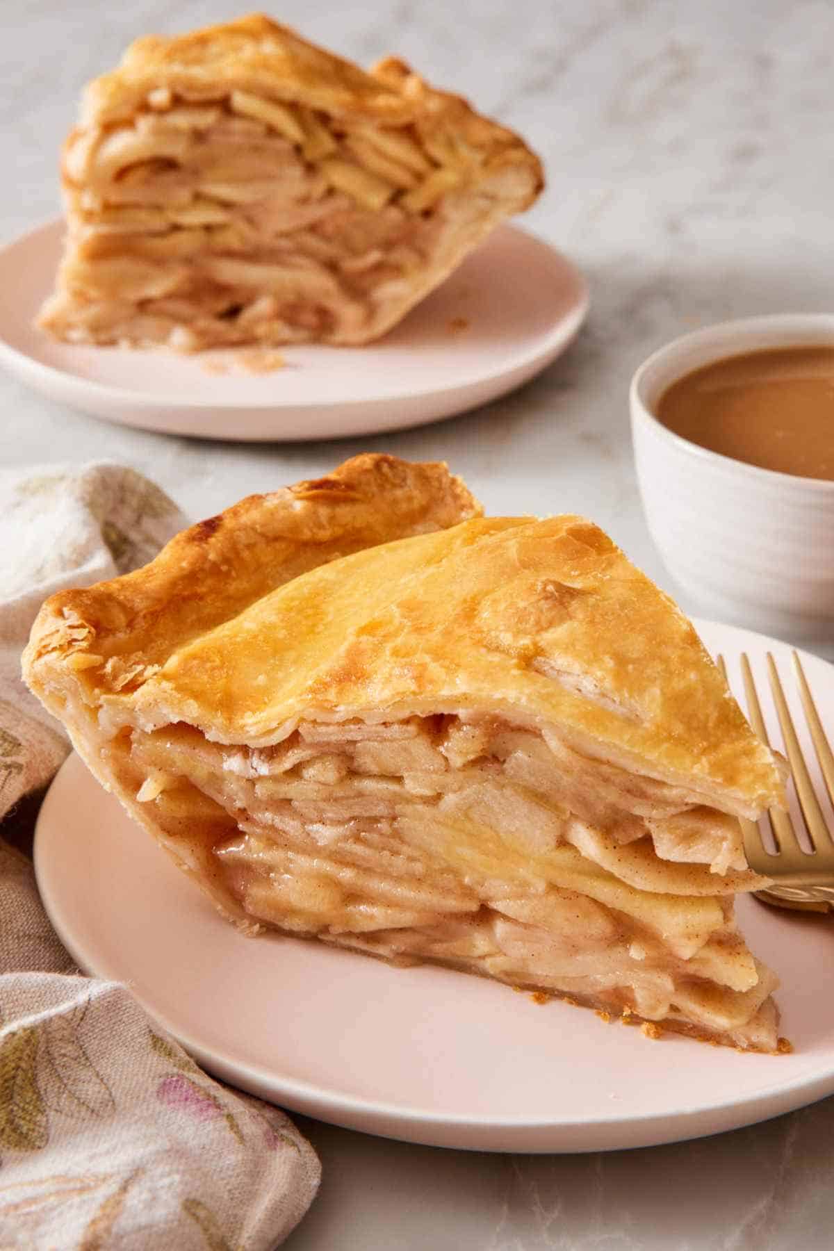 A slice of apple pie on a plate with a cup of coffee. Another slice is in the background out of focus.