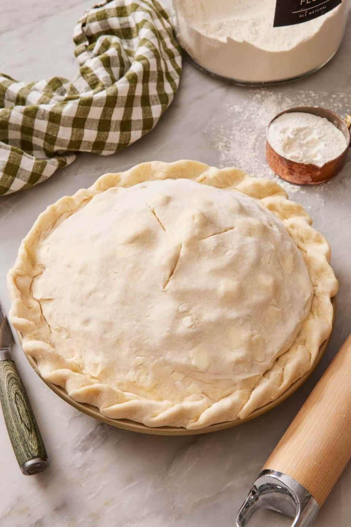 An unbaked double crust pie on a counter with a table linen and measuring cup of flour behind it.