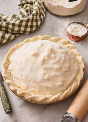 An unbaked double crust pie on a counter with a table linen and measuring cup of flour behind it.