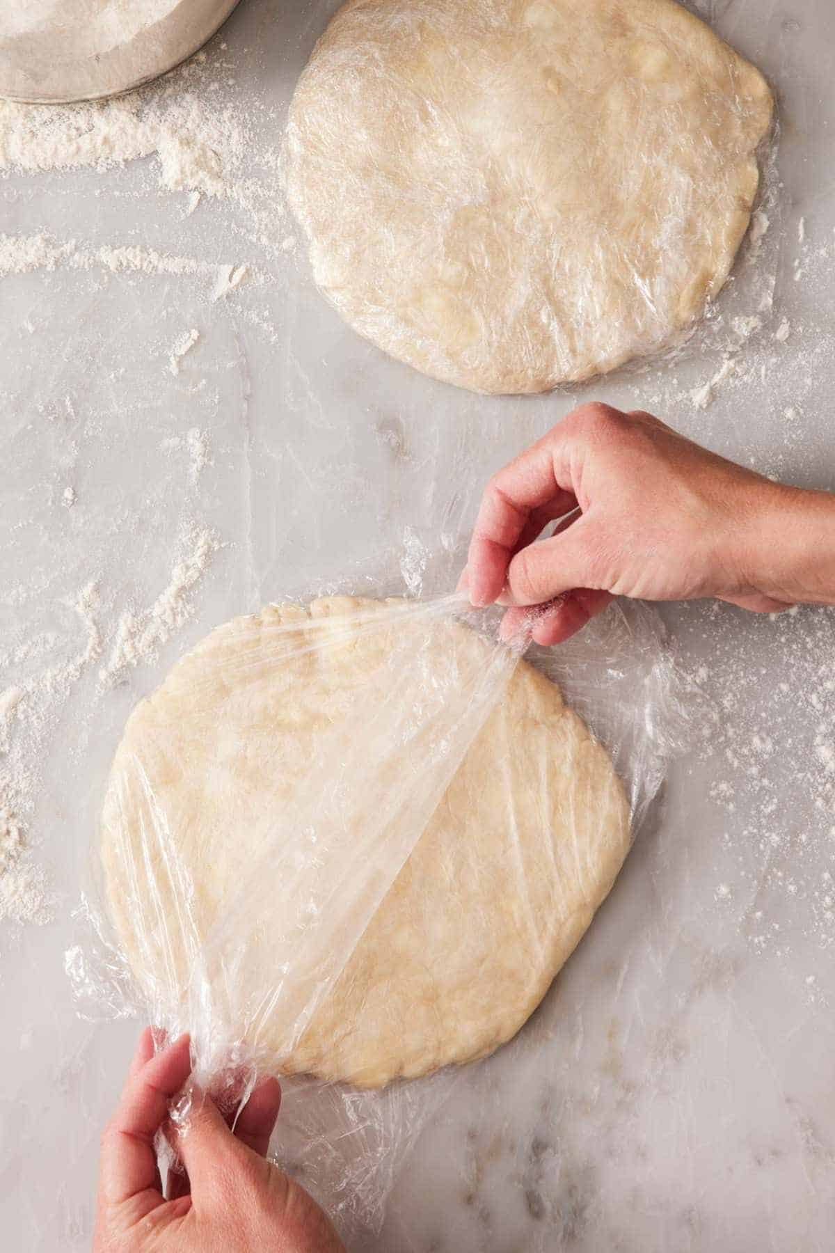 A disc of pie crust dough being wrapped with plastic wrap alongside another already wrapped disc.