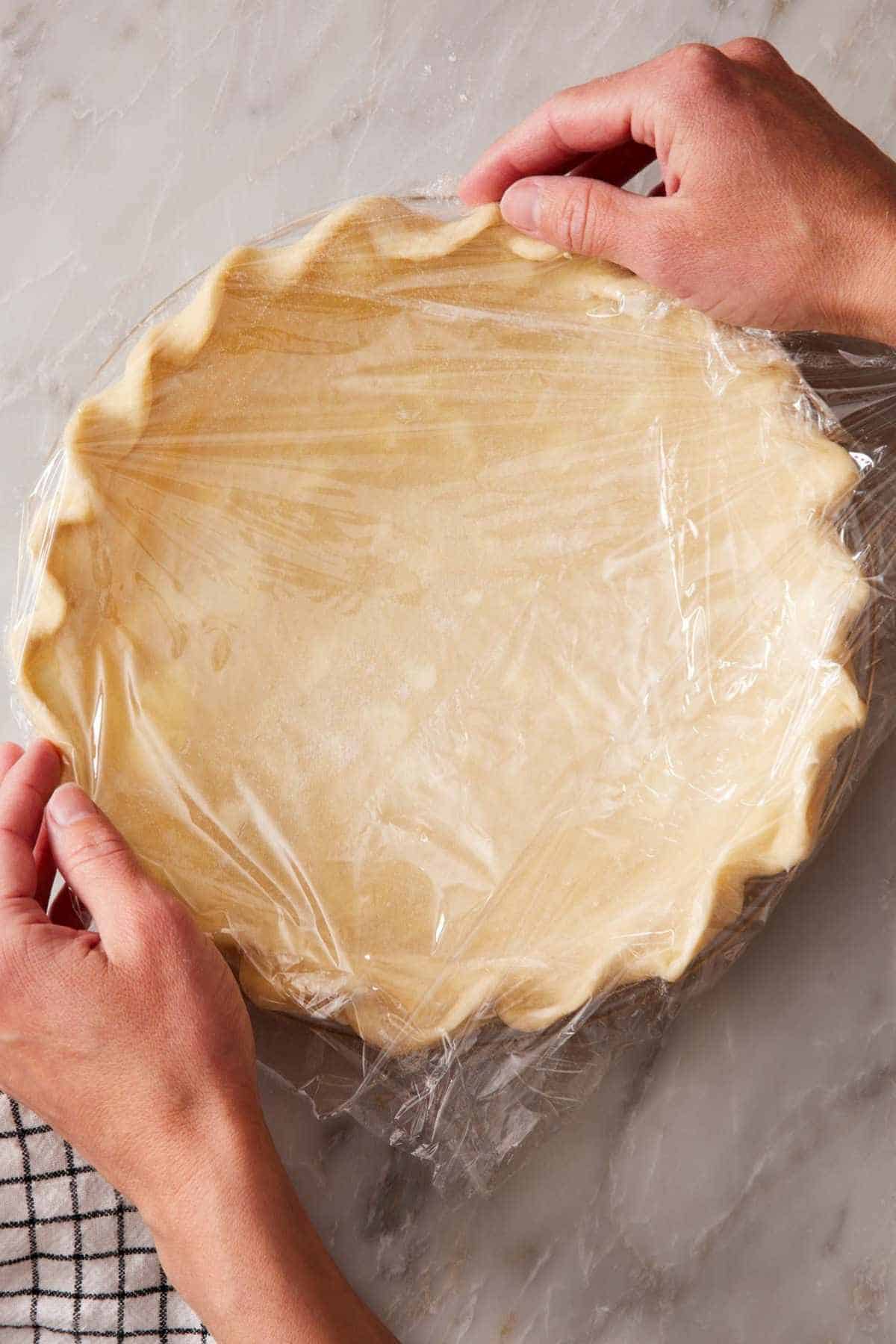 A crimped pie crust in a pie pan wrapped in plastic wrap for freezing.
