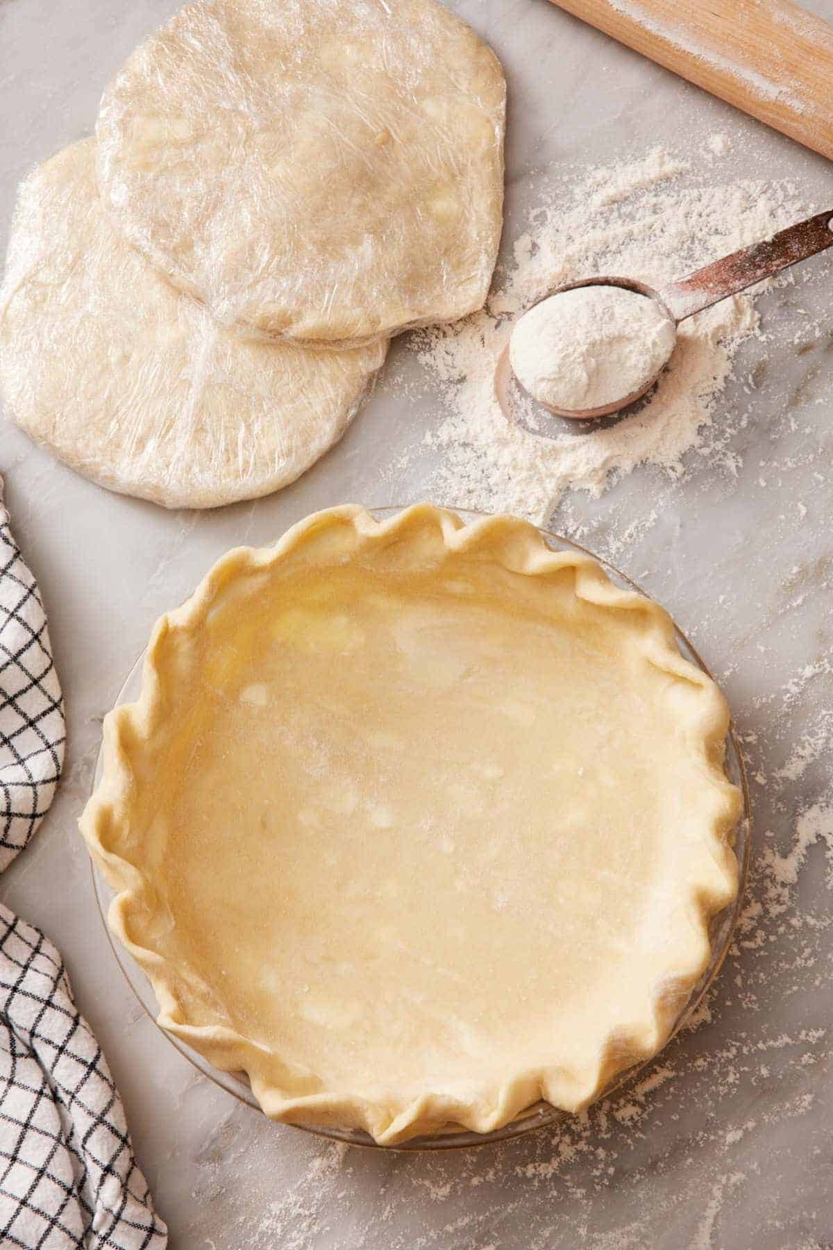 A crimped pie crust in a pie dish alongside two plastic-wrapped discs of pie dough.