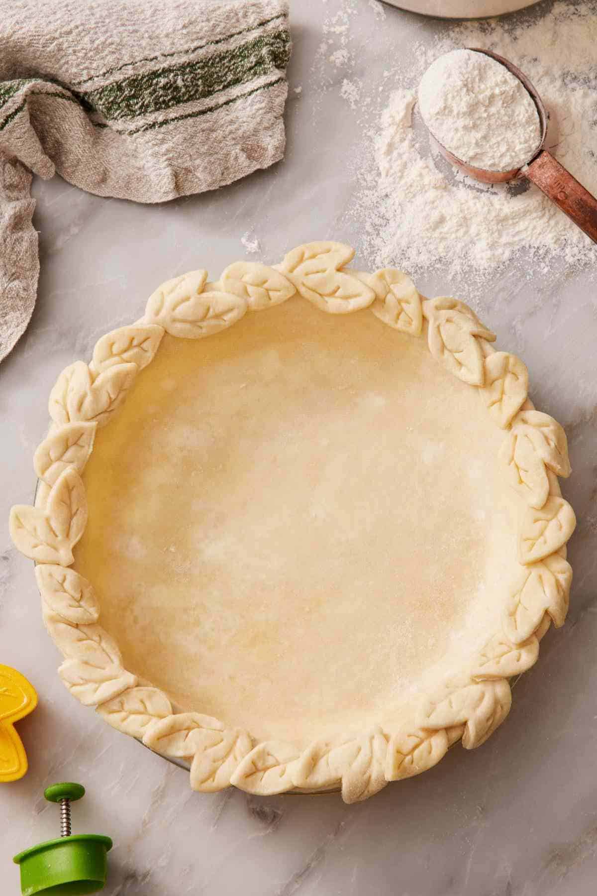 A pie crust that has a decorative crimp on a countertop with baking supplies almost out of frame surrounding it.