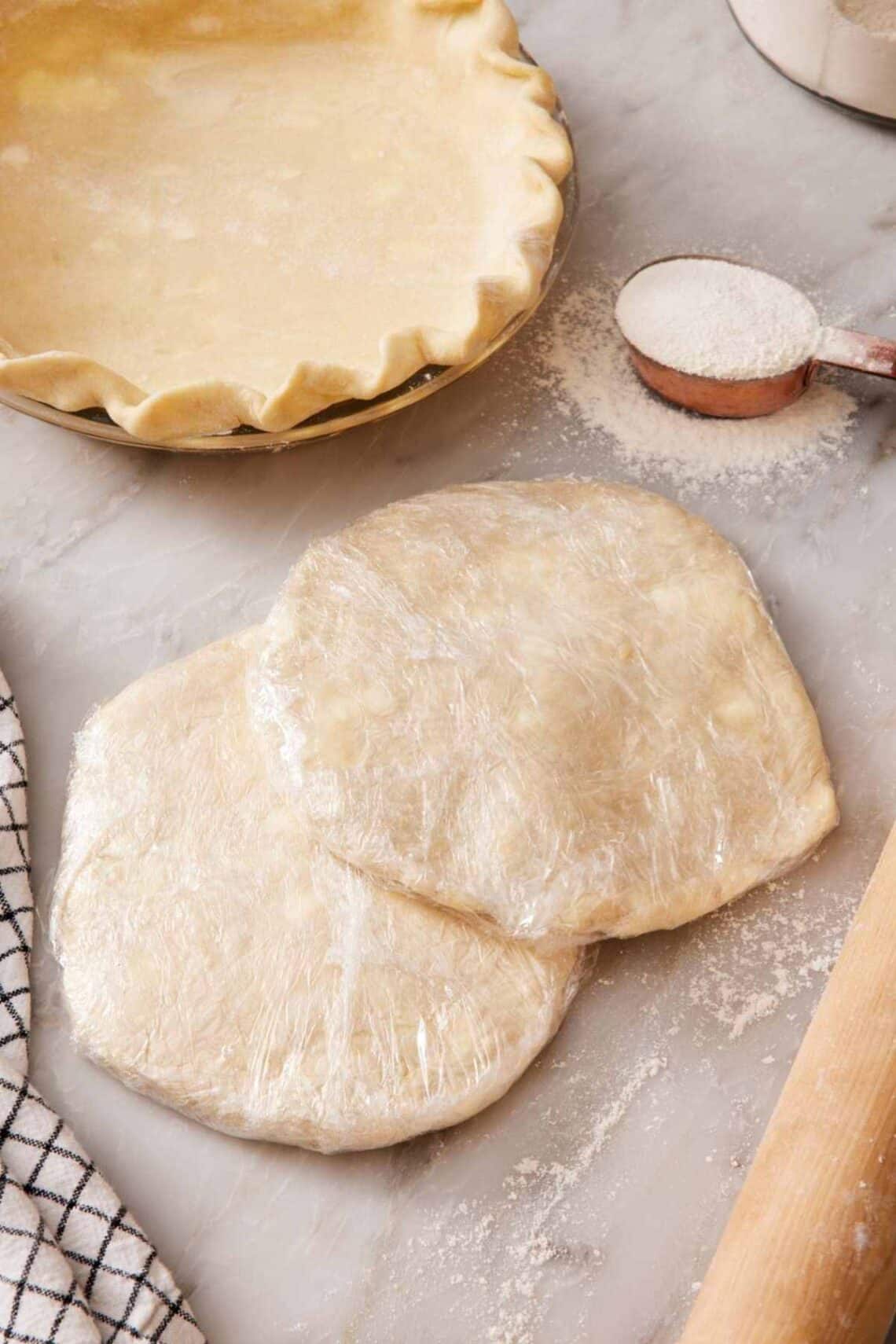 Two discs of wrapped pie dough partially stacked, with a measuring cup of flour and a prepared pie crust in a pie dish in the background.