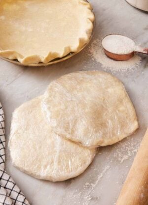 Two discs of wrapped pie dough partially stacked, with a measuring cup of flour and a prepared pie crust in a pie dish in the background.
