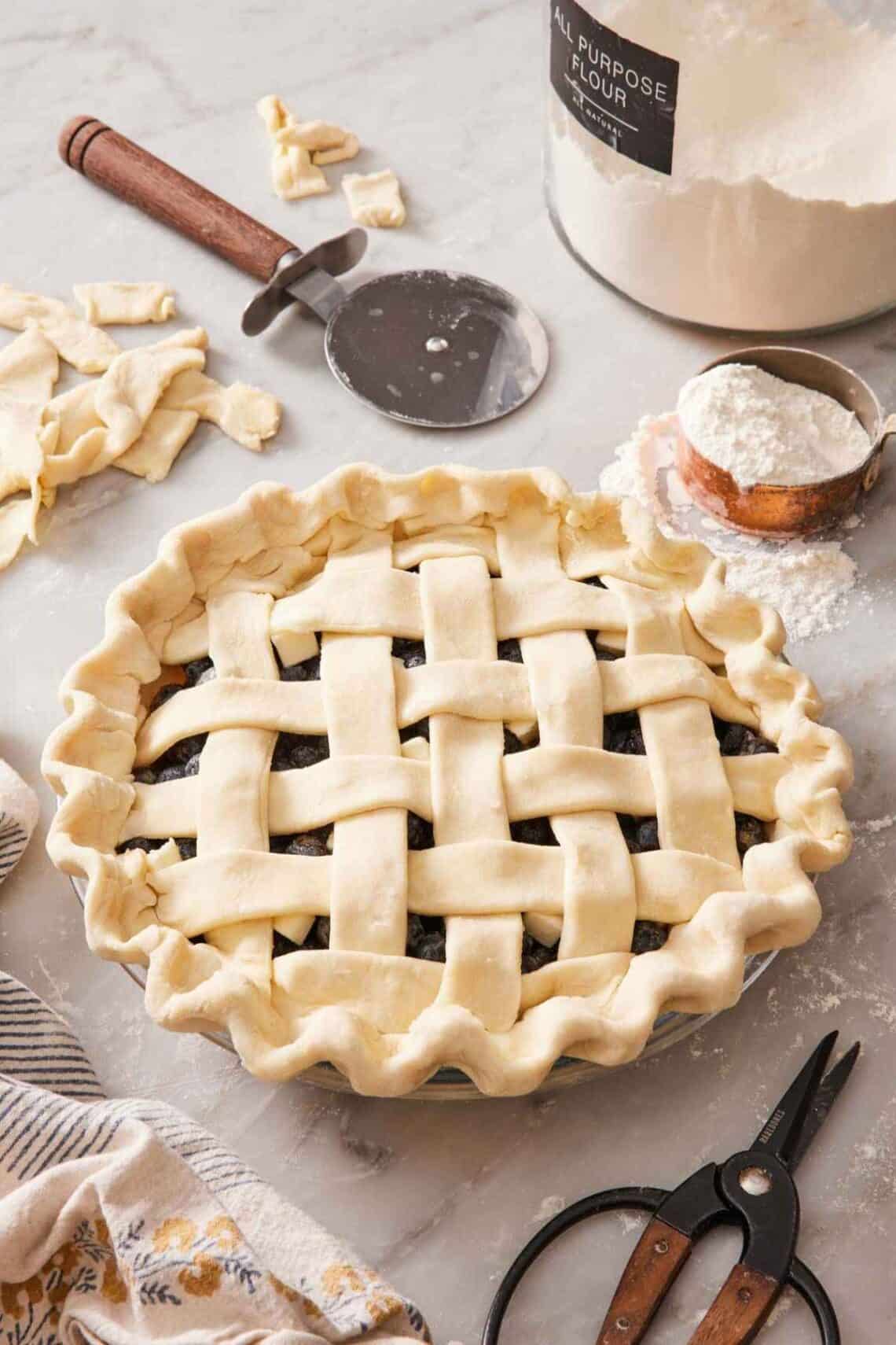 A pie with a lattice top on a counter with baking tools surrounding it.