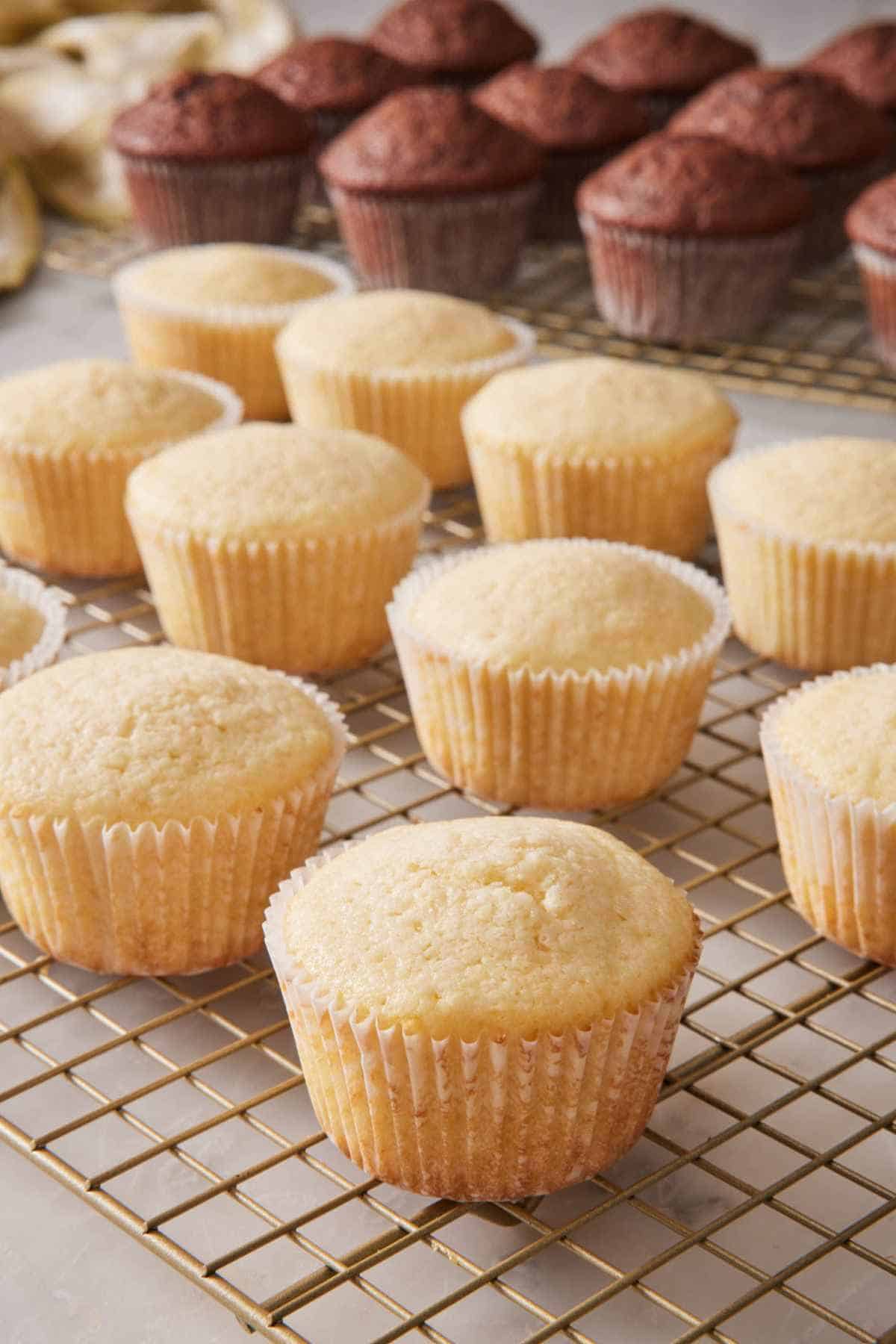 Vanilla cupcakes on a wire rack, with chocolate cupcakes on a cooling rack behind them.
