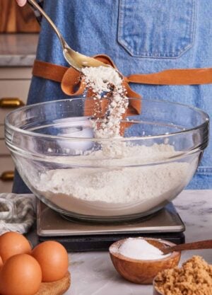 Flour spooned into a bowl containing more flour on a kitchen scale. More baking ingredients in the foreground.